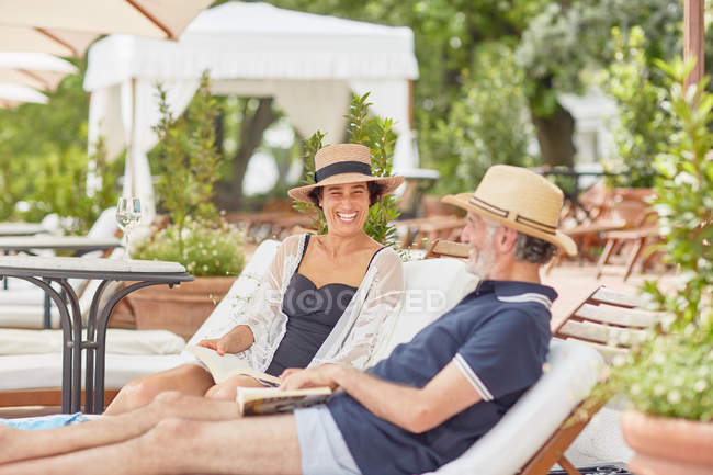Couples d'âge mûr lisant des livres, se relaxant sur des chaises longues au bord de la piscine — Photo de stock