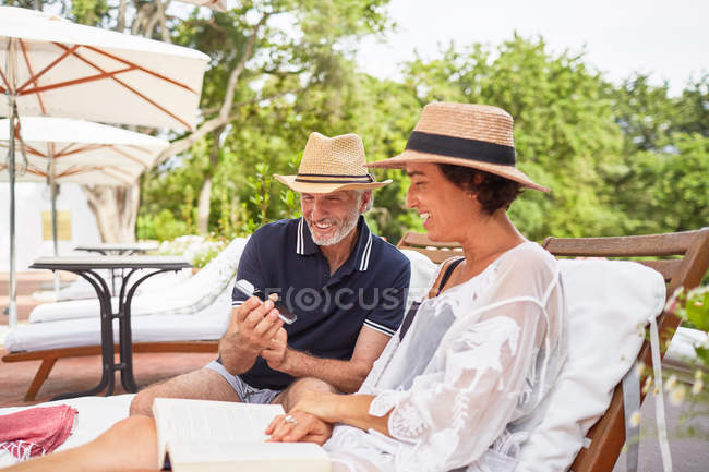 Couple d'âge mûr avec téléphone intelligent relaxant sur des chaises longues au bord de la piscine du complexe — Photo de stock