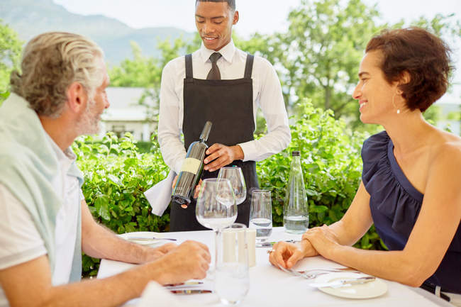 Weinbegleiter zeigt reifem Paar Weinflasche beim Essen auf der Terrasse — Stockfoto