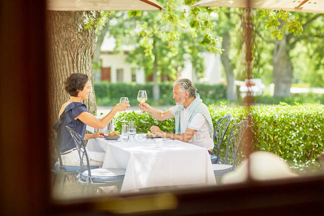 Pareja madura brindando copas de vino en la mesa del patio del resort - foto de stock