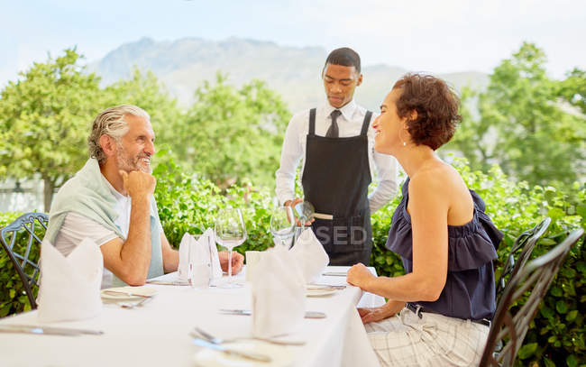 Vinho mordomo derramando vinho para casal maduro jantando na mesa do pátio — Fotografia de Stock