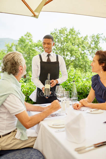 Camarero de vino mostrando botella de vino a pareja cenando en la mesa del patio - foto de stock