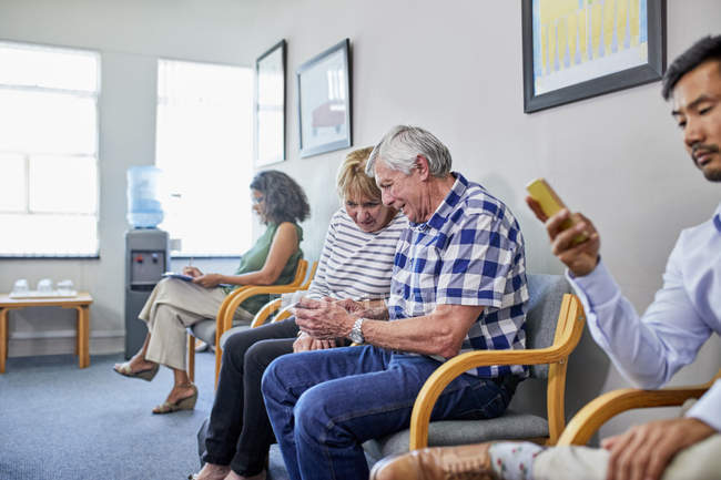 Casal sênior usando telefone inteligente na sala de espera clínica — Fotografia de Stock