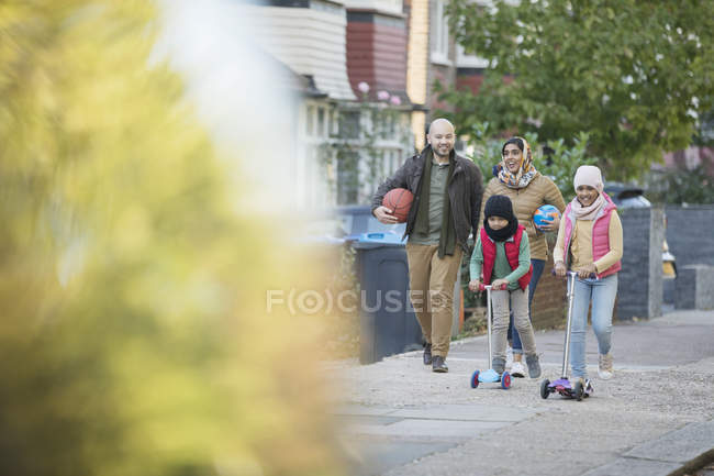 Familia musulmana caminando y montando scooters en la acera del barrio - foto de stock