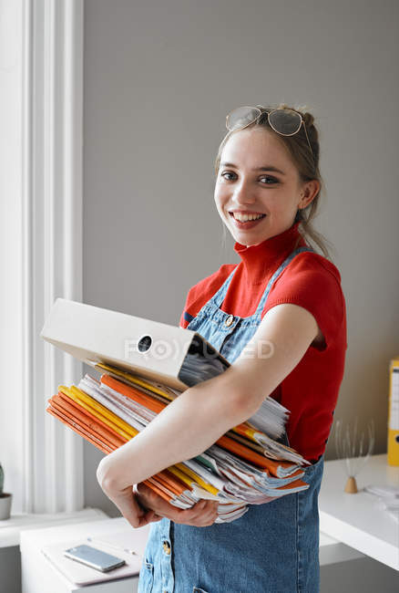 Retrato confiado joven estudiante universitaria llevando pila de libros y carpeta - foto de stock