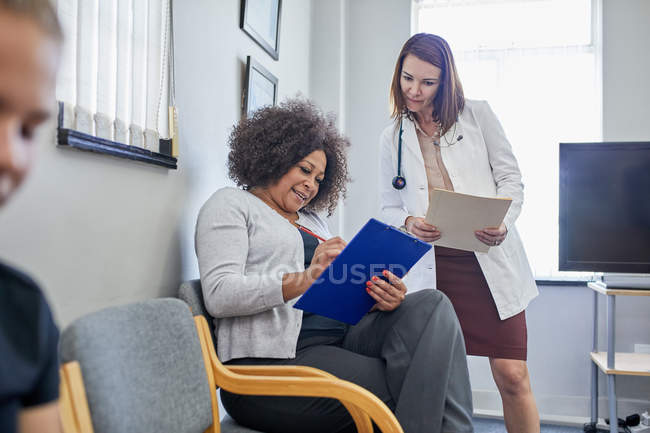 Doctora y paciente discutiendo papeleo en sala de espera clínica - foto de stock