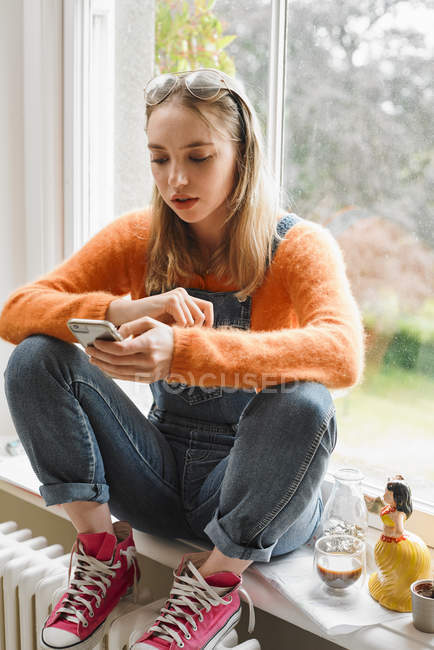 Молодая женщина смс со смартфоном в окне — стоковое фото