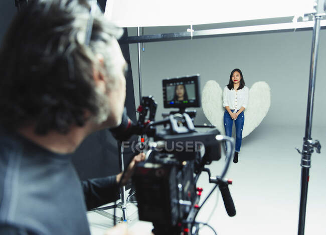 Photographer behind camera photographing young woman wearing angel wings during photo shoot in studio — Stock Photo