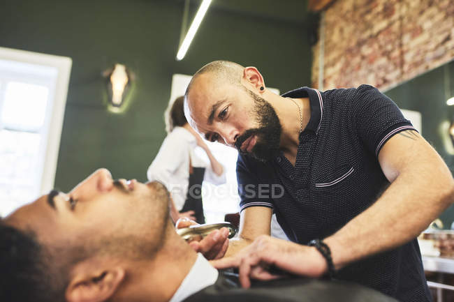 Peluquero masculino enfocado que da al cliente un afeitado en la barbería - foto de stock