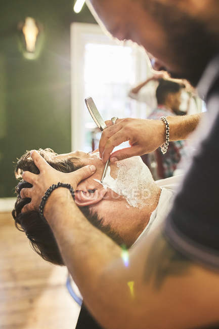 Peluquero macho que da al cliente un afeitado en la barbería - foto de stock