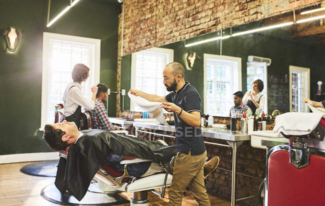 Peluquero macho preparándose para la cara de vapor del cliente antes de afeitarse en la barbería - foto de stock