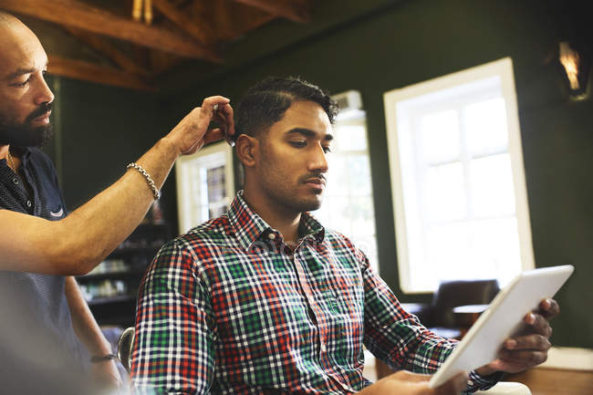 Hombre usando tableta digital mientras recibe corte de pelo en la barbería - foto de stock