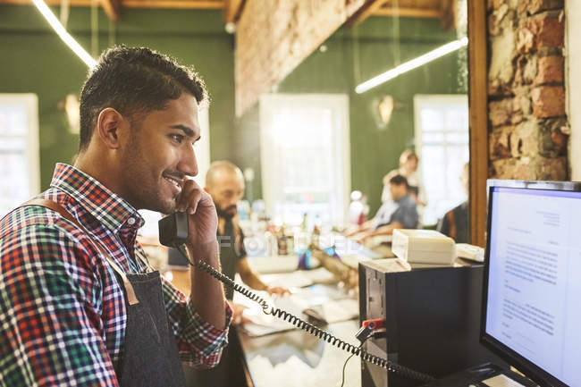 Male barber talking on telephone at computer in barbershop — Stock Photo