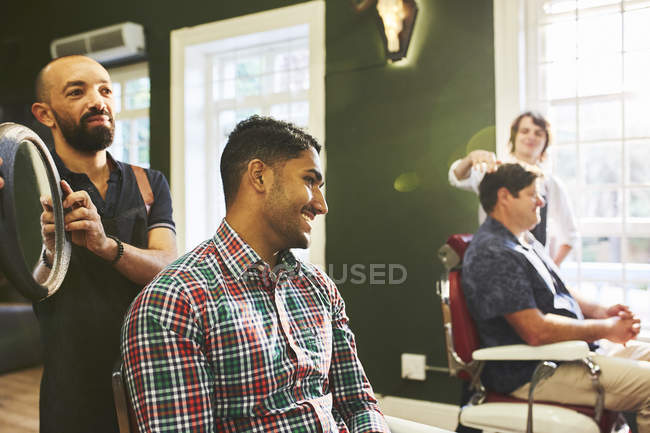 Peluquero masculino que sostiene el espejo para el cliente sonriente en la barbería - foto de stock