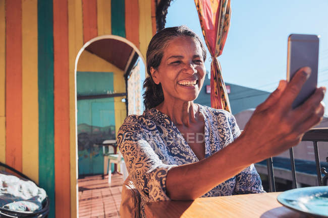 Усміхаючись, щаслива жінка бере селфі зі смартфоном на сонячному патіо — стокове фото
