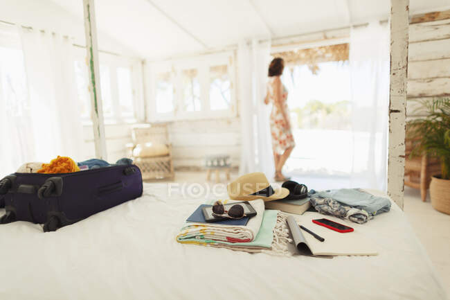 Mujer parada en la puerta de la cabaña de la playa más allá de la maleta y pertenencias en la cama - foto de stock