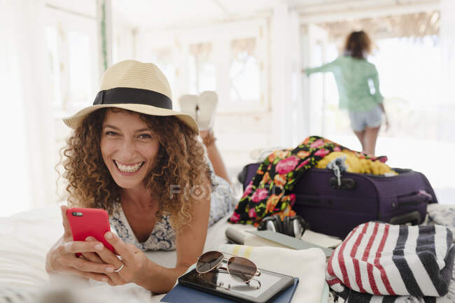 Portrait happy young woman using smart phone, unpacking suitcase in bedroom — Stock Photo