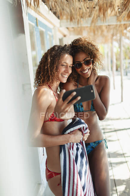 Felice giovani donne amiche in bikini prendendo selfie con smart phone sulla spiaggia capanna patio — Foto stock