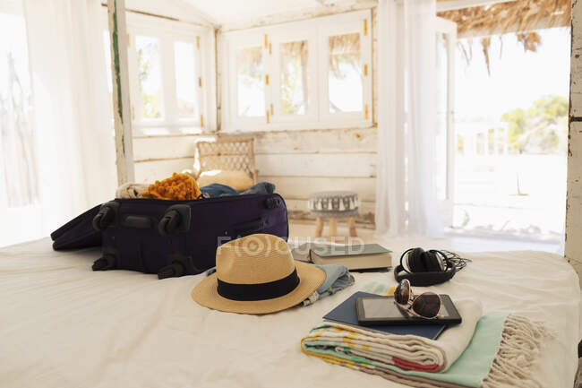 Валіза, сонячний капелюх, сонцезахисні окуляри, книга та цифровий планшет на ліжку пляжного будинку — стокове фото