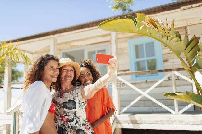 Mãe feliz e filhas adultas tomando selfie fora da cabana de praia ensolarada — Fotografia de Stock
