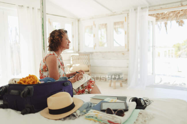 Serene woman writing in journal next to suitcase in beach hut bedroom — Stock Photo