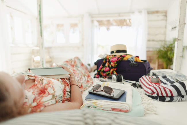 Woman with book relaxing on bed next to suitcase in sunny beach hut bedroom — Stock Photo