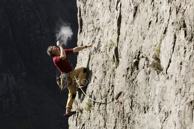 Männlicher Bergsteiger erklettert Felswand, blickt auf und bläst Kreide auf die Hände — Stockfoto