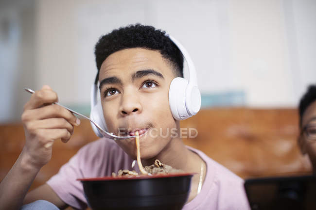Мальчик-подросток в наушниках ест лапшу дома — стоковое фото