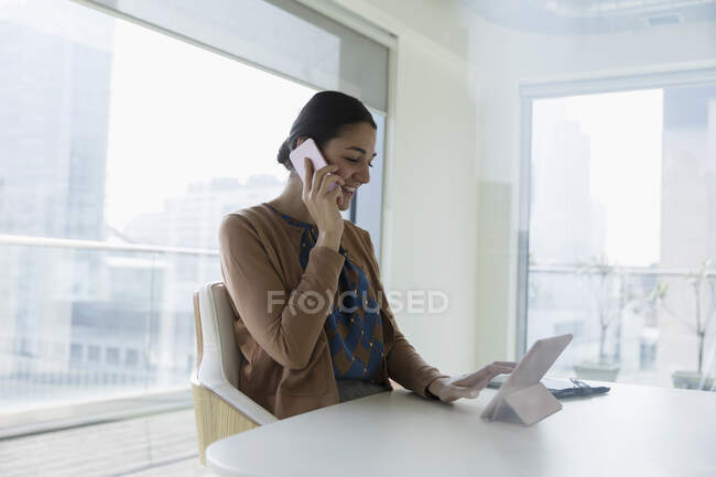 Smiling businesswoman talking on smart phone and using digital tablet in office — Stock Photo