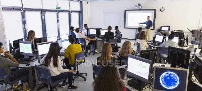 Estudiantes de secundaria usando computadoras y viendo al profesor en la pantalla de proyección en el aula - foto de stock