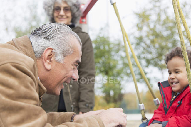 Grandfather playing with grandson on swing at playground — Stock Photo