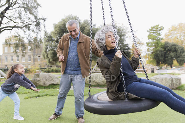 Abuelos juguetones y nieta jugando en swing en el parque - foto de stock