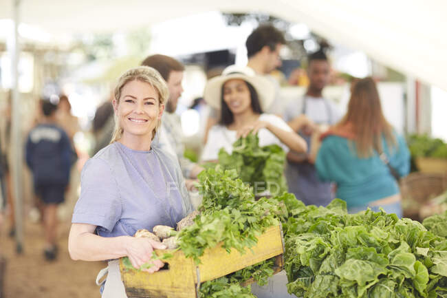 Retrato sonriente, mujer confiada trabajando, llevando cajón de verduras en el mercado de agricultores - foto de stock