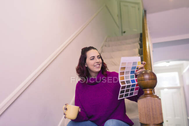 Улыбающаяся женщина перекрашивает, смотрит на образцы краски на лестнице — стоковое фото