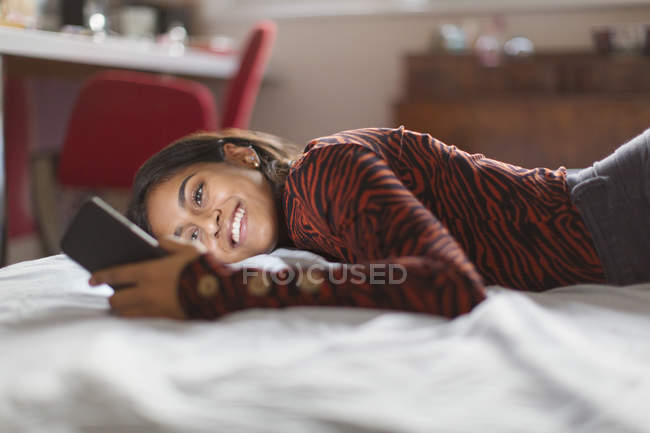 Улыбающаяся девочка-подросток, использующая смартфон на кровати — стоковое фото