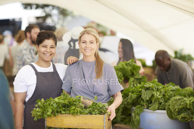 Retrato sonriente de las trabajadoras con cajón de verduras en el mercado de agricultores - foto de stock