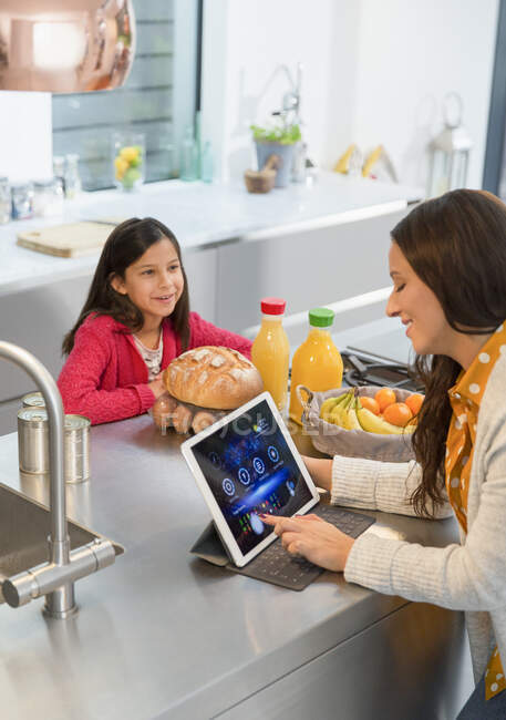 Дочка дивиться, як мати використовує цифровий планшет на кухні — стокове фото