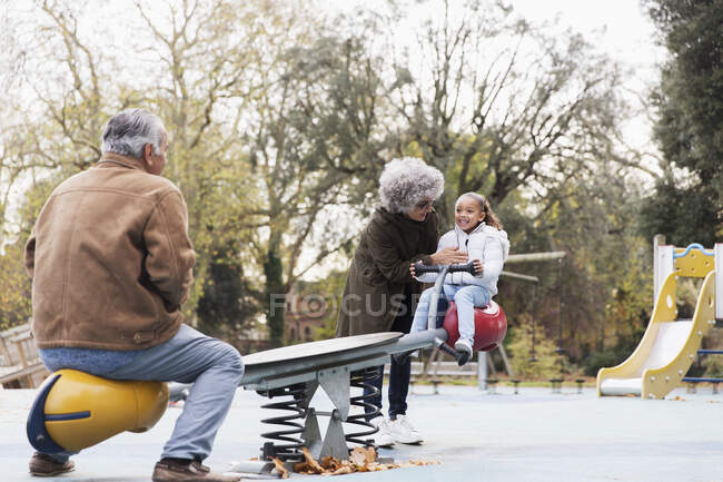 Бабушка с дедушкой играют с внучкой на качелях на детской площадке — стоковое фото