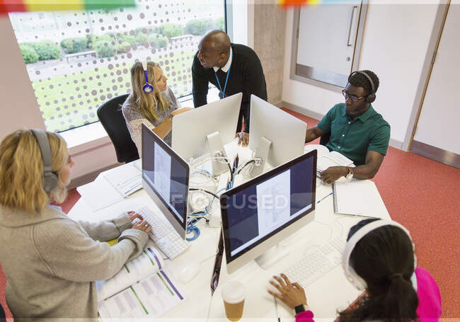 Instrutor universitário comunitário ajudando os alunos com fones de ouvido usando computadores no laboratório de informática — Fotografia de Stock