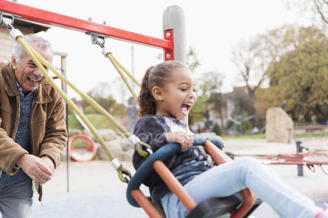 Playful grandfather pushing granddaughter on playground swing — Stock Photo