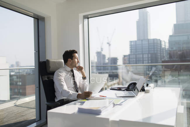 Pensativo hombre de negocios con papeleo en oficina soleada, moderna, urbana - foto de stock
