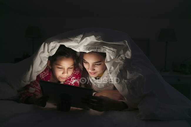 Hermanas viendo película en tableta digital bajo manta en dormitorio oscuro - foto de stock
