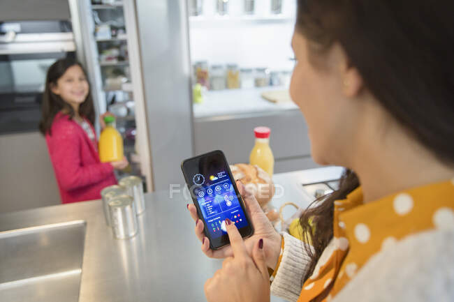 Mother using smart phone app to track groceries in refrigerator, watching daughter — Stock Photo