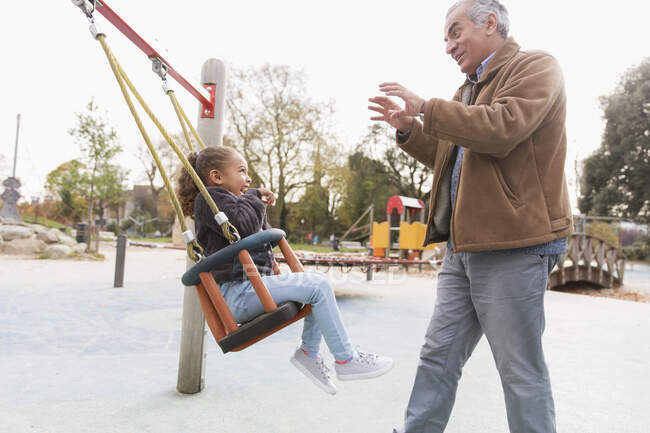Grandfather pushing granddaughter on playground swing — Stock Photo