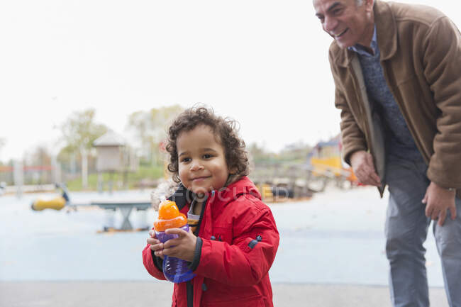 Дедушка смотрит на внука малыша с кружкой на детской площадке — стоковое фото