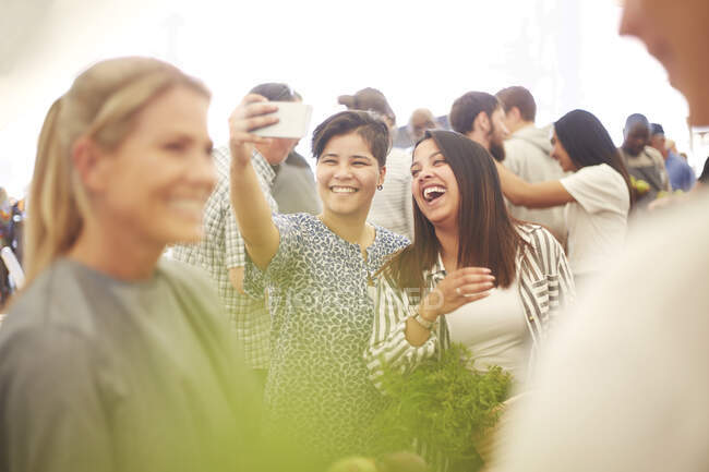 Feliz, entusiasta pareja de lesbianas tomando selfie en el mercado de agricultores - foto de stock