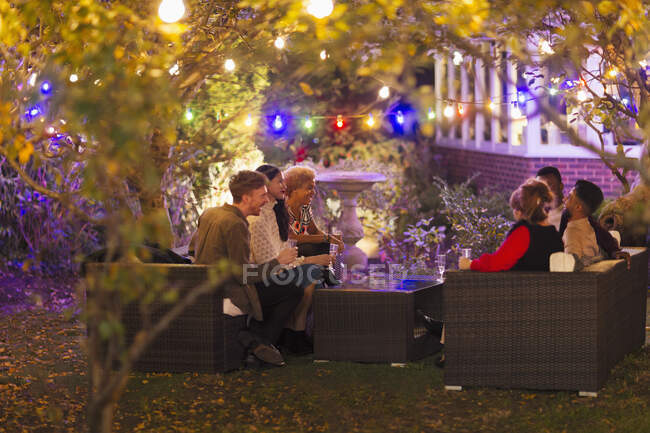 Amigos hablando y bebiendo bajo los árboles con luces de cuerda en la fiesta del jardín - foto de stock