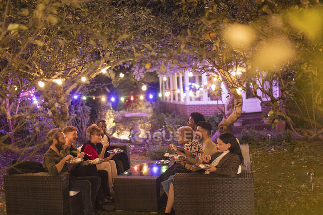 Amici che parlano e mangiano dessert sotto gli alberi con luci di spago alla festa in giardino — Foto stock