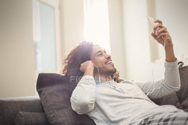 Sonriente joven relajante, escuchando música con teléfono inteligente y auriculares en el sofá - foto de stock