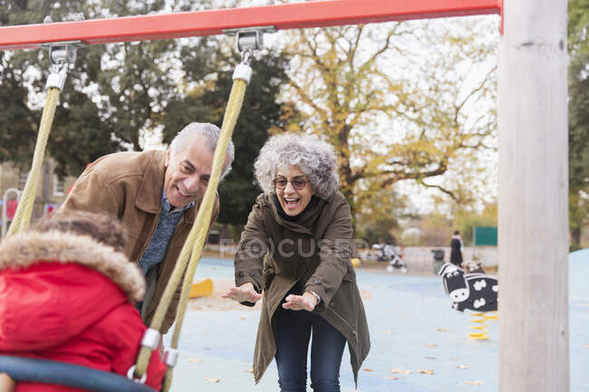 Grand-parents ludiques poussant petit-fils tout-petit sur la balançoire à aire de jeux — Photo de stock